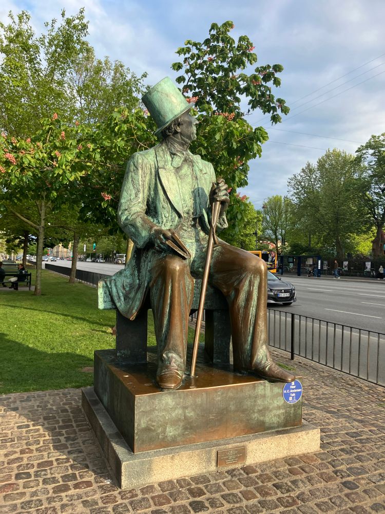 A bronze statue of Hans Christian Anderson in Copenhagen. He is sitting, wearing a top hat, has a book on his lap, and a walking stick in his hand 