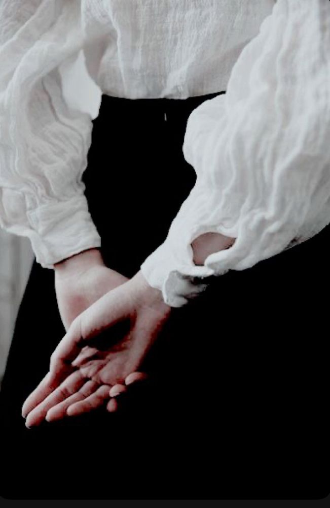 A photograph of a woman in historical clothing from behind. She’s wearing a white, wide sleeved, Edwardian shirt and a black skirt. 