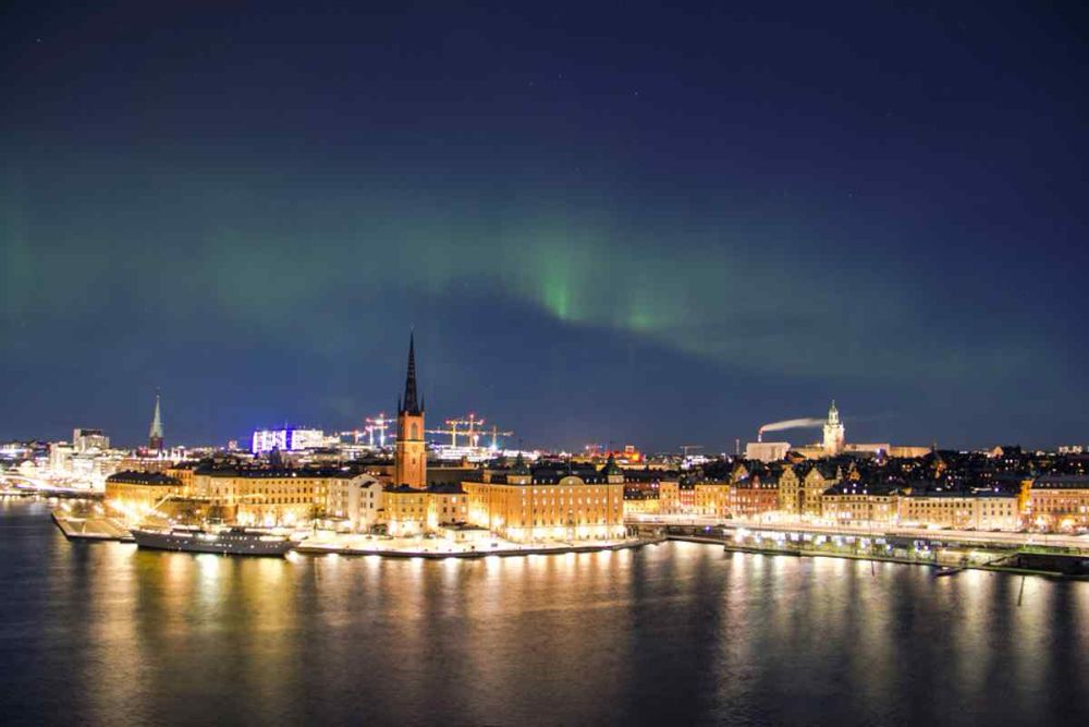 Image of the aurora borealis over Stockholm city. The city is brightly lit in warm tones. The aurora is green against a blue sky, but no stars are visible 