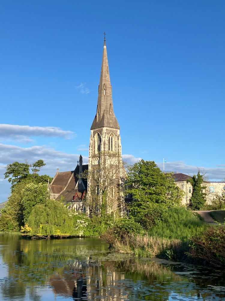 Photograph of St. Alban’s church in Copenhagen. In the forefront is a moat with lilly pads. The river bank is lined with green trees including willows. The church is behind the willows. The church is from 1887 and has a spire and stained glass windows 