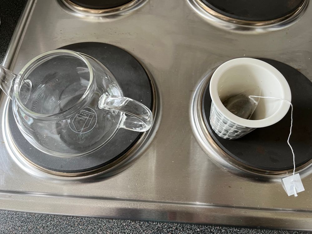 A glass teapot on one rubber electric hob, with it’s ceramic tea filter sitting on the hob next to it. The ceramic filter has a grey pattern on the outside and a black teabag with a string inside.