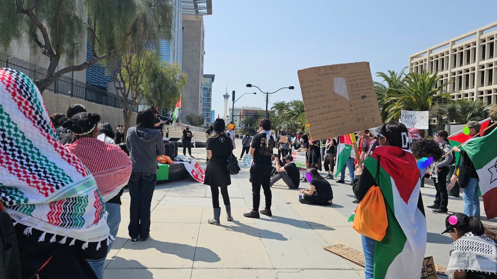 Backs of people, some wearing kufiyas and Palestinian flags with signs