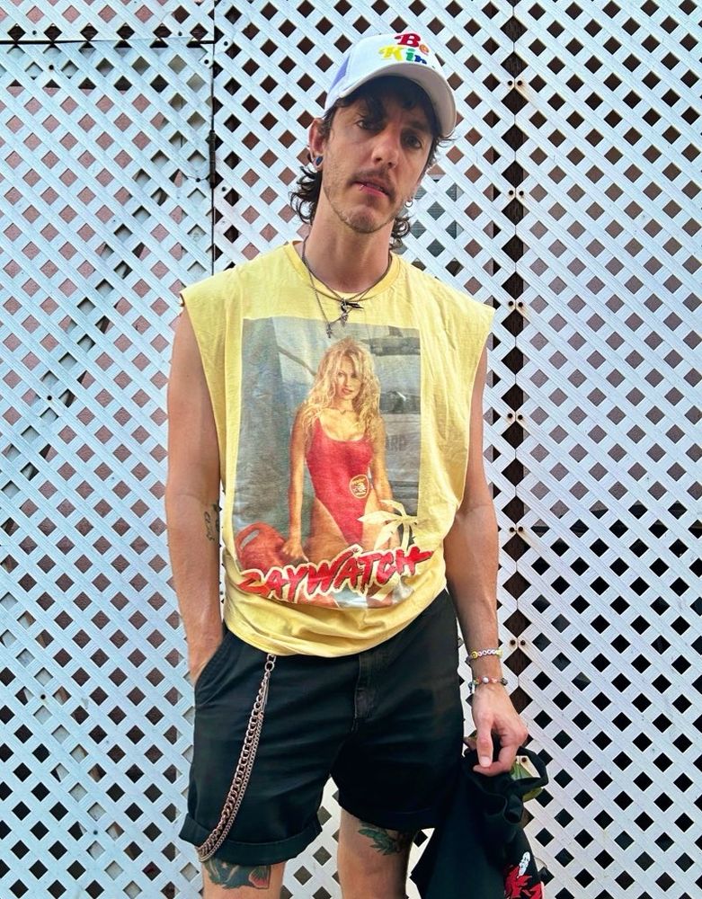 Kasey in a yellow cutoff that says “Baywatch” featuring a photo of Pamela Anderson in her signature red swimsuit from the show. Kasey wears a white baseball cap + black shorts.