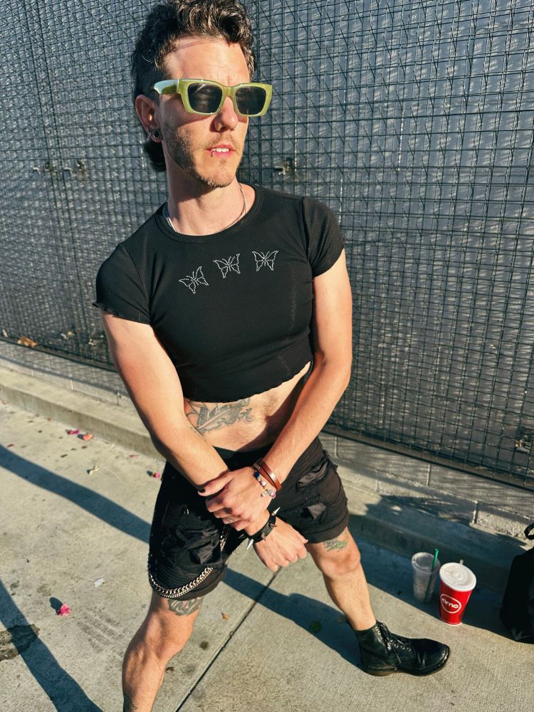 Kasey poses in a black crop top with 3 rhinestone butterflies across the chest. He wears green sunglasses, black shorts, black boots. 