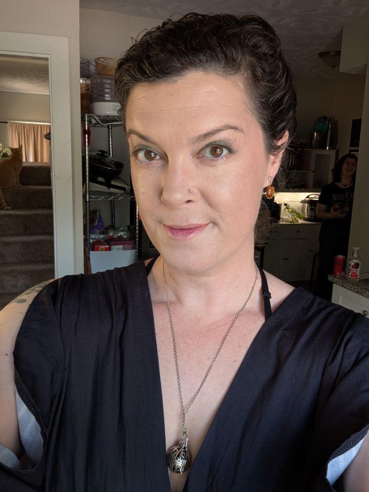 Selfie of a white brunette femme with their hair swept in an up-do, pale makeup, and Sakura style lips wearing a flowy black dress and a silver pomander necklace 