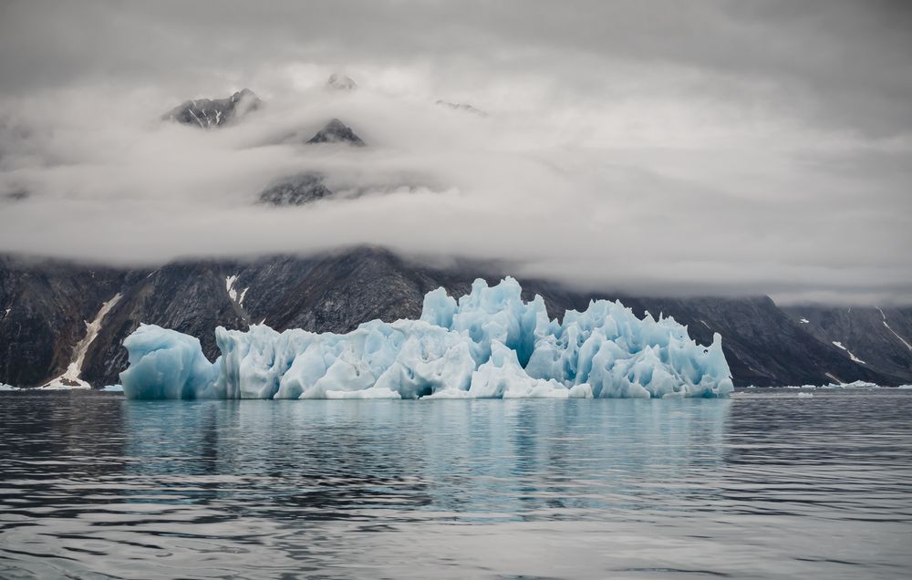 A blue iceberg with jagged peaks reflects in the calm water. In the background the peaks of a mountain poke through a low, dense cloude cover. Photo from East Greenland by Lori Ryerson.