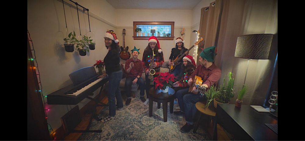 Screen capture from video showing a family of six wearing festive hats and playing instruments in a living room. Camera tricks were used to make it look like three people are clones.