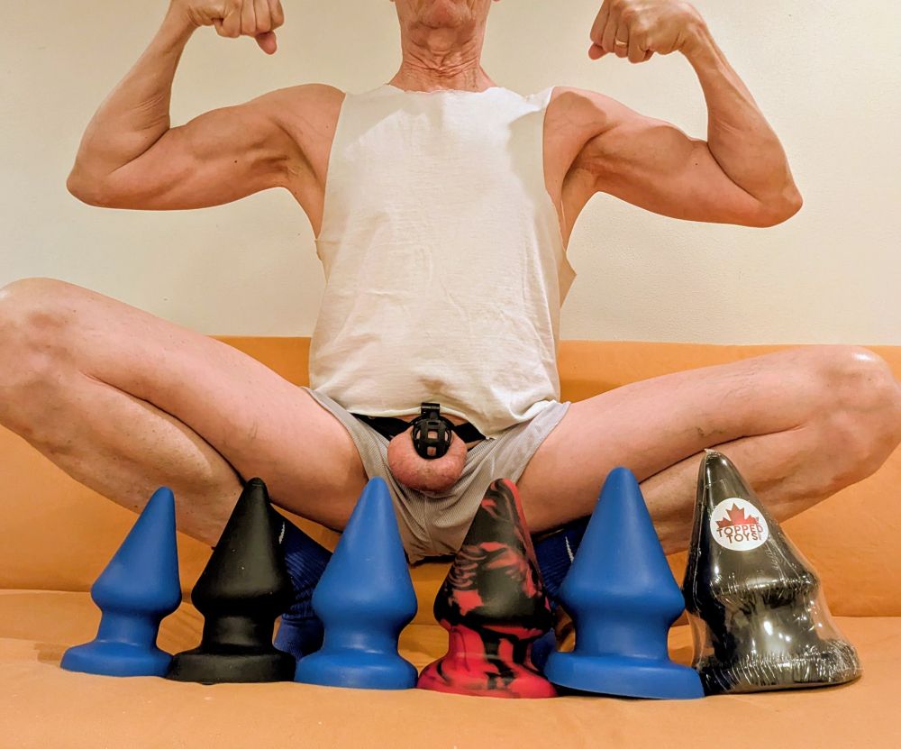 A lineup of 6 Grip plugs on an orange couch, from the 96 on the left, progressing to the 150 (still wrapped in plastic) on the right. 
I'm squatted above the toys, knees out to either side, my caged dicklet exposed, doing a two arm bicep pose. I'm wearing a white muscle tee shirt and very skimpy and slutty grey short shorts. 