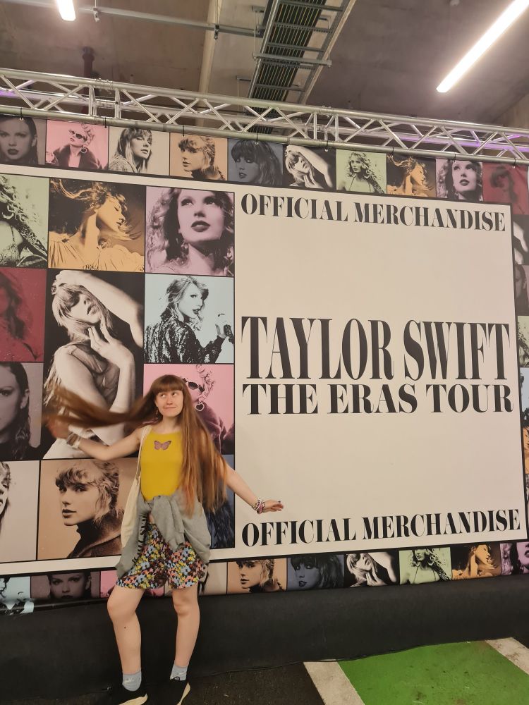 Flora, a woman with long blonde hair standing in front of a wall that says 'Official Merchandise, Taylor Swift the Eras Tour' with eras pictures all over it