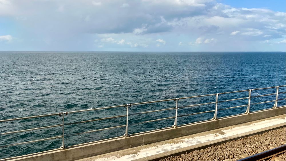 Blick aus dem Zug. Unten erkennt man eine Schiene, dann ein Geländer, darüber das Meer mit einigen Wolken 