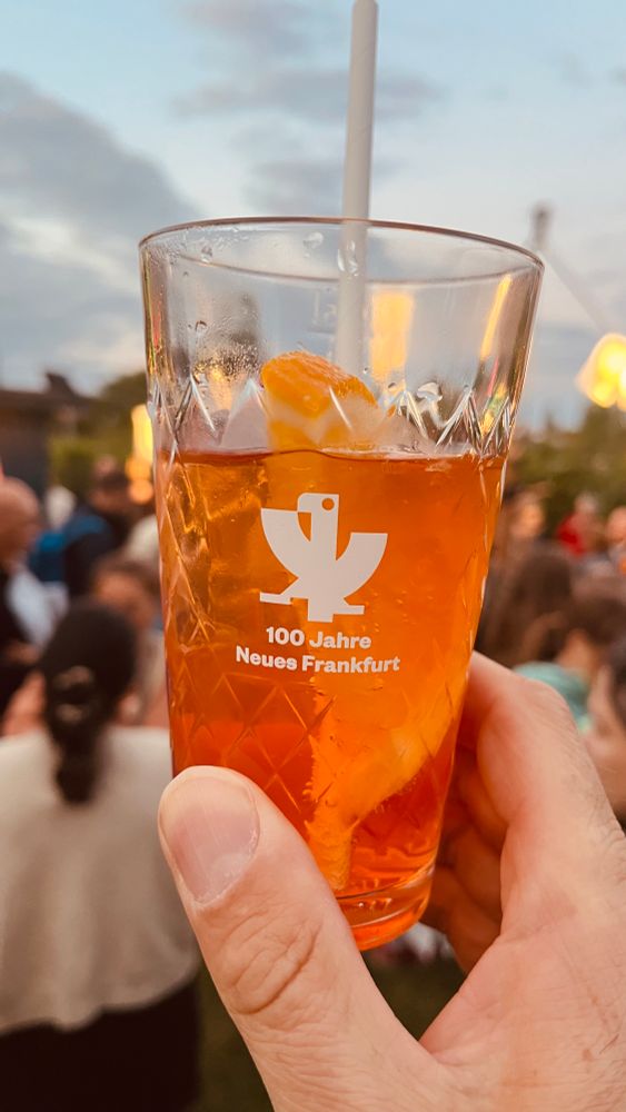 Ein Geripptes mit orange farbener Flüssigkeit. Auf dem Glas das Wappen von Frankfurt aus der Zeit des neuen Frankfurt. Darunter steht „100 Jahre Neues Frankfurt „
