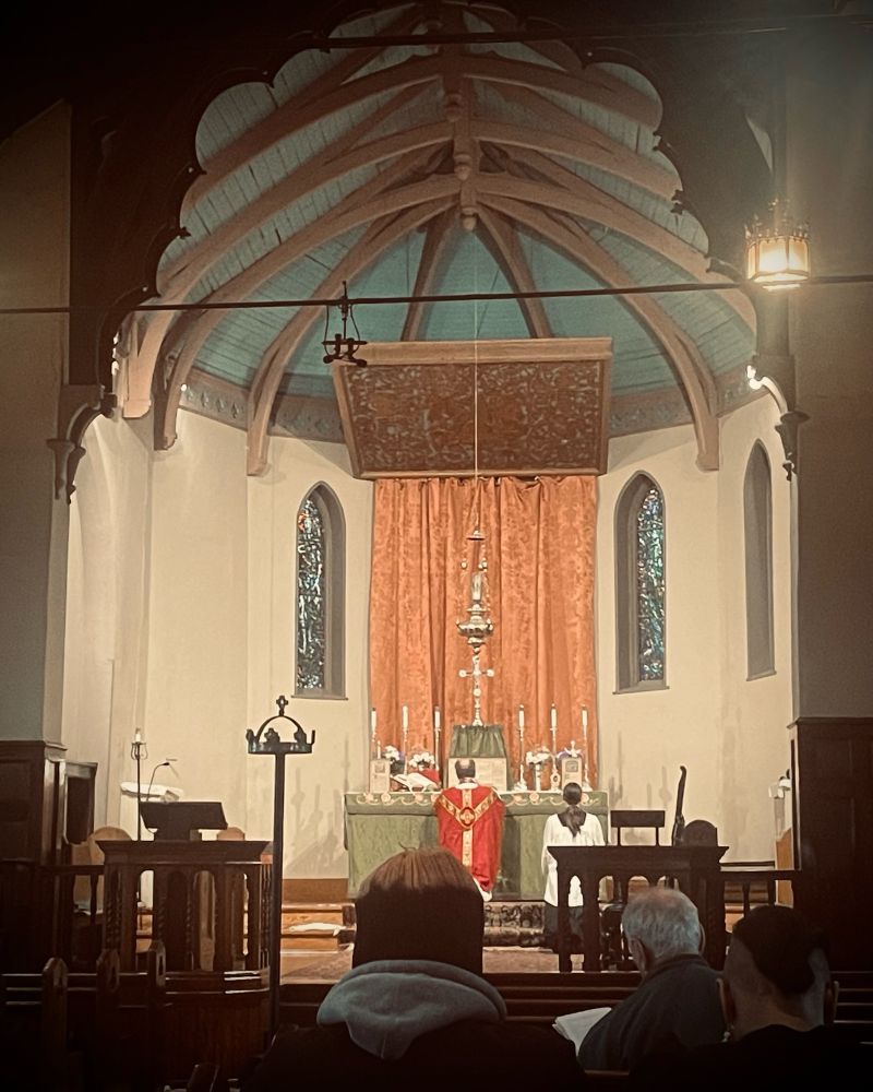 Picture of the chancel of S. Bartholomew’s church from the back pews, with a few congregants, and server and priest at the altar. Green paraments, pink dossal, red vestments.
