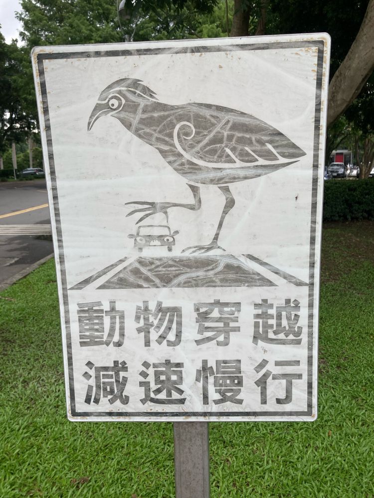 A faded sign that reads, in traditional Chinese, “animal crossing, reduce speed” and has a picture of an enormous bird in the foreground with a tiny car in the background, looks like the bird is about to pluck the car up and eat it.