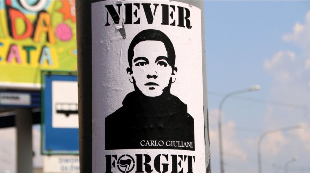 Das Bild zeigt ein Sticker, der an einem Pfosten angebracht ist. Auf dem Sticker ist ein schwarz-weißes Porträt eines jungen Mannes zu sehen. Über dem Porträt steht in großen, schwarzen Buchstaben "NEVER" und darunter "FORGET". Der Buchstabe O bei FORGET ist ein Antifa Logo. Unter dem Bild ist der Name "CARLO GIULIANI" abgedruckt.

Im Hintergrund ist eine unscharfe städtische Umgebung zu erkennen, mit einem bunten Wandbild oder Werbeplakat und einigen Straßenschildern sowie einem blauen Himmel.