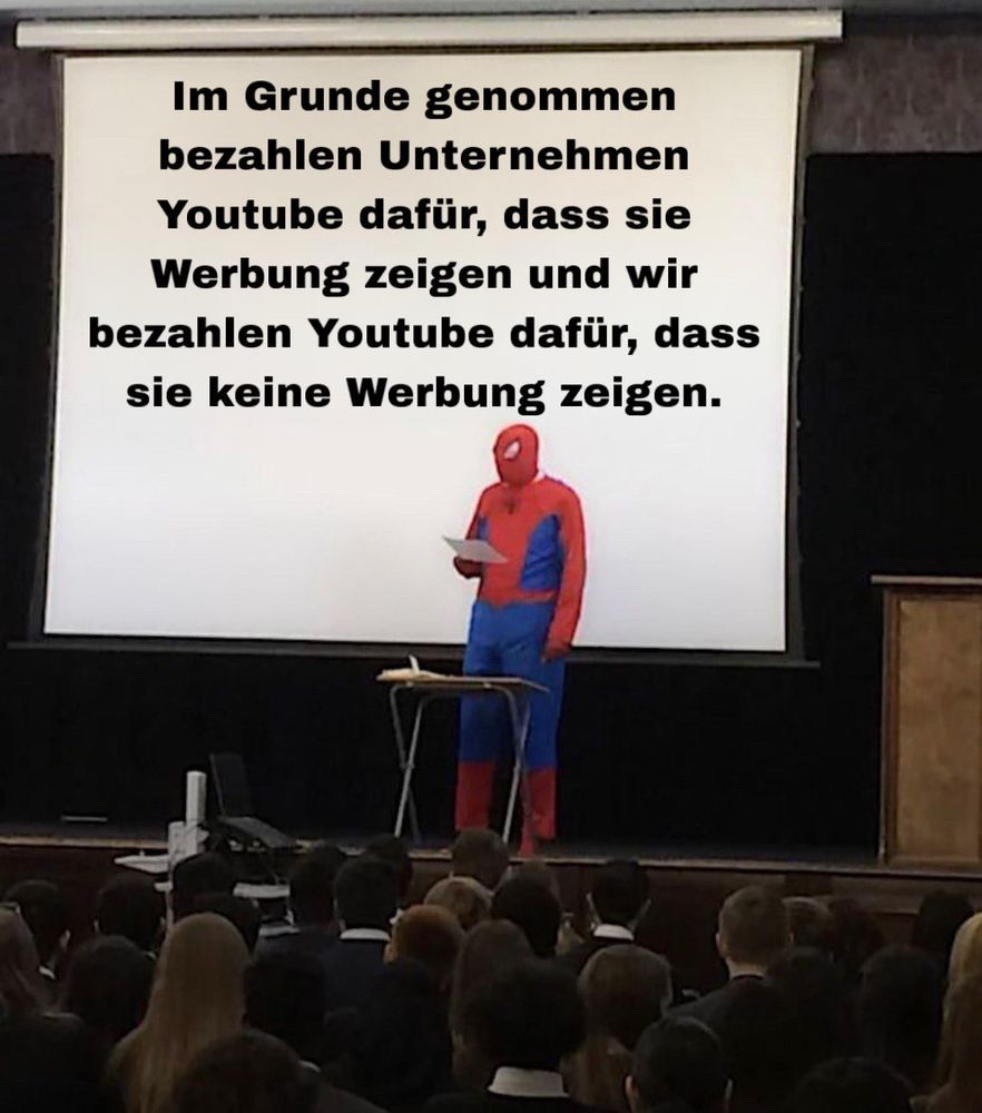 Eine Person im Spiderman Kostüm steht auf der Bühne vor Publikum und hält einen Zettel in der Hand.
Hinter der Person ist eine Leinwand auf der der Text „Im Grunde genommen bezahlen Unternehmen
Youtube dafür, dass sie Werbung zeigen, und wir bezahlen Youtube dafür, dass sie keine Werbung zeigen.“ projiziert wird.  