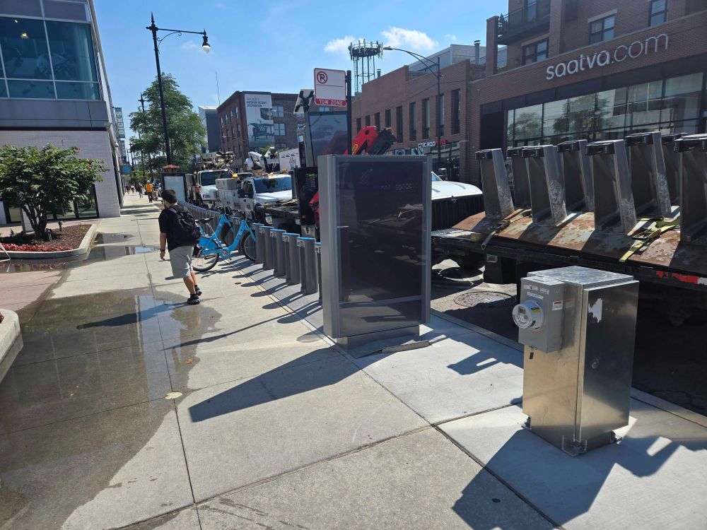 Side view of the new Divvy station installation showing the full dock line, a pedestrian walking by, and nearby plaza space.