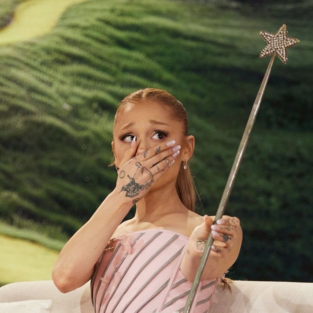 Ariana Grande emotional in shock with hand covering mouth while holding up old Glinda wand from original movie. 