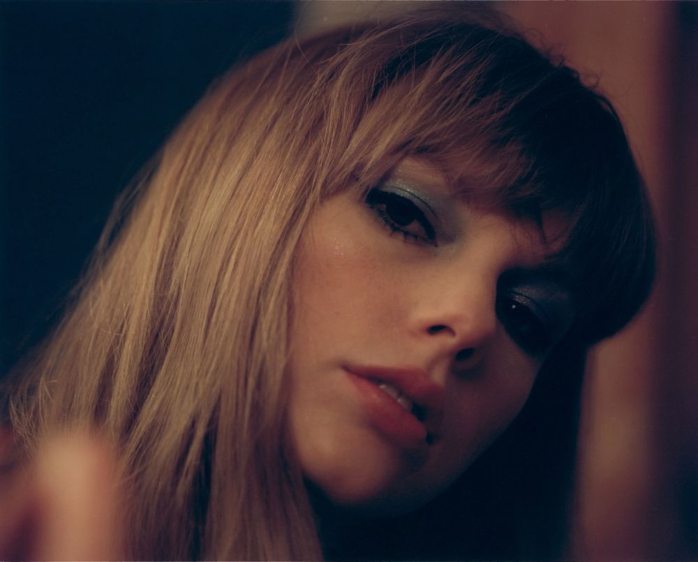 Taylor Swift for the ‘Midnights’ photoshoot wearing blue sparkly eyeshadow and a light red lipstick while standing in a gloomy room and looking at the camera sleepily. 