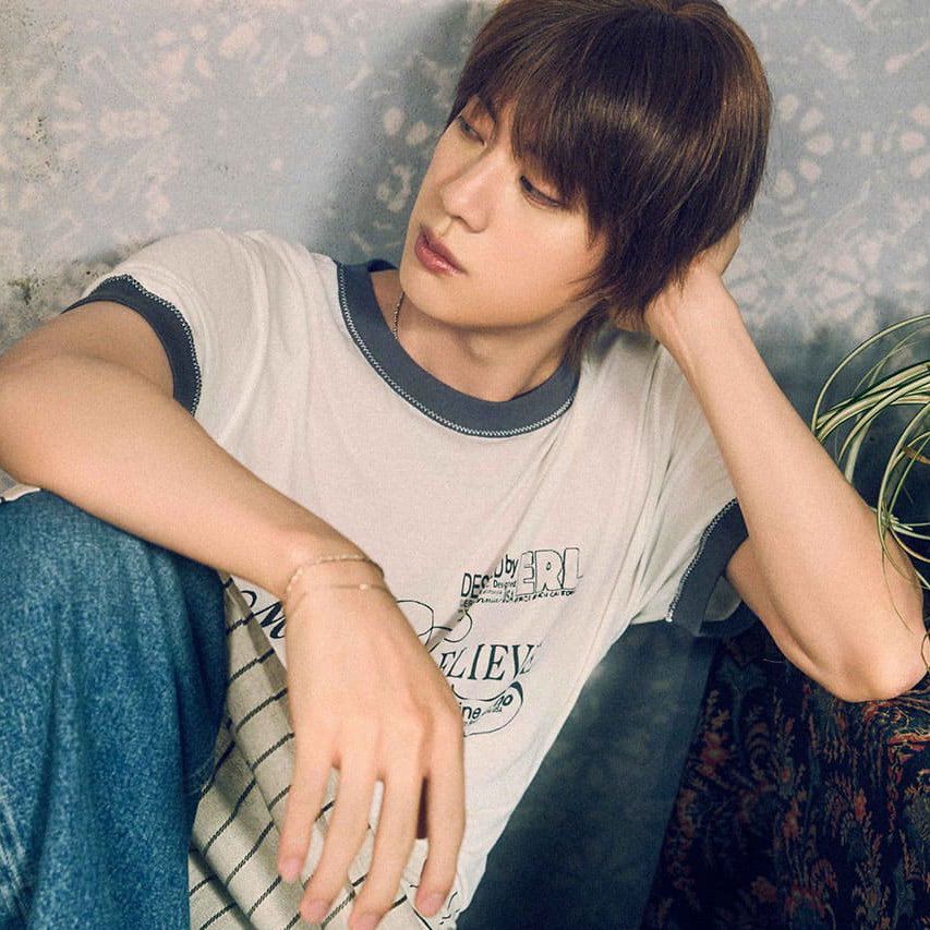 JIN wearing a white shirt and jeans sitting down against a wall with his hand on his head looking to his right. 