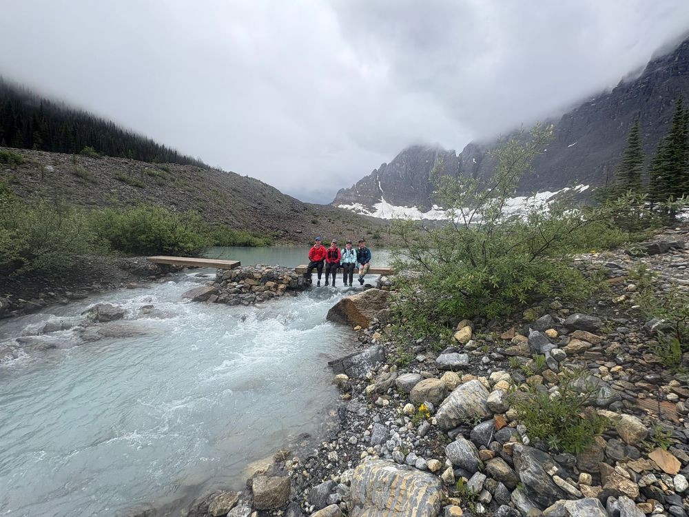 Trail runners sitting on bridge over a glacier fed stream with mountains in the background 