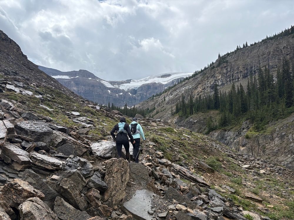 Two runners navigating boulders of a rock slide to find the trail on the other side. A mountain valley in the background. 