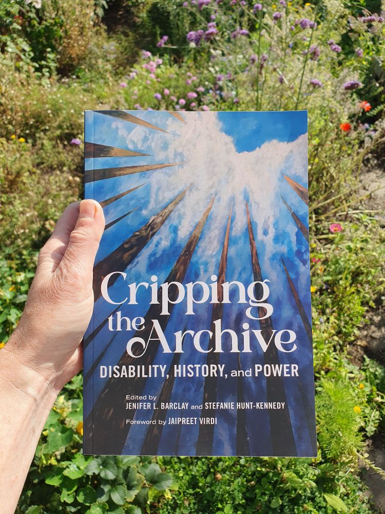 A hand holding a book in a sunny garden. The book has a blue cover with striking grey shards, and the title Cripping the Archive: Disability, history and power