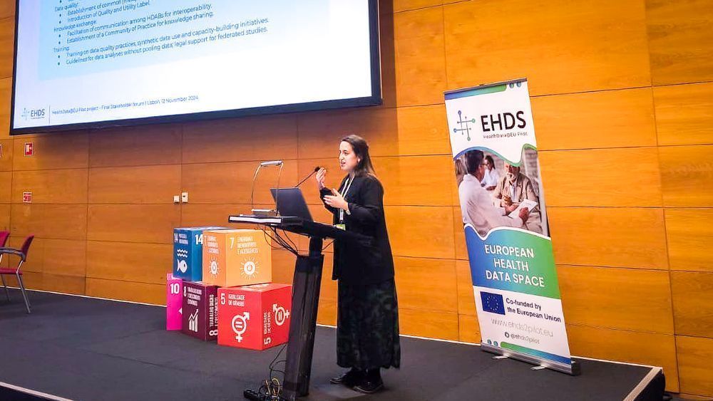 A dark-haired woman, dressed in black, gives a talk at a stage of the European Public Health Conference. Next to her, a roll-up highlights the EHDS, the European health data space.