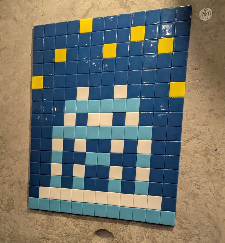 Photo d'une mosaïque de l'artiste "Space Invader", sur un mur en béton gris. C'est une grille de 12×15 carreaux, représentant un extra-terrestre très pixelisé en bleu clair et blanc sur fond bleu marine avec quelques pixels jaunes au dessus