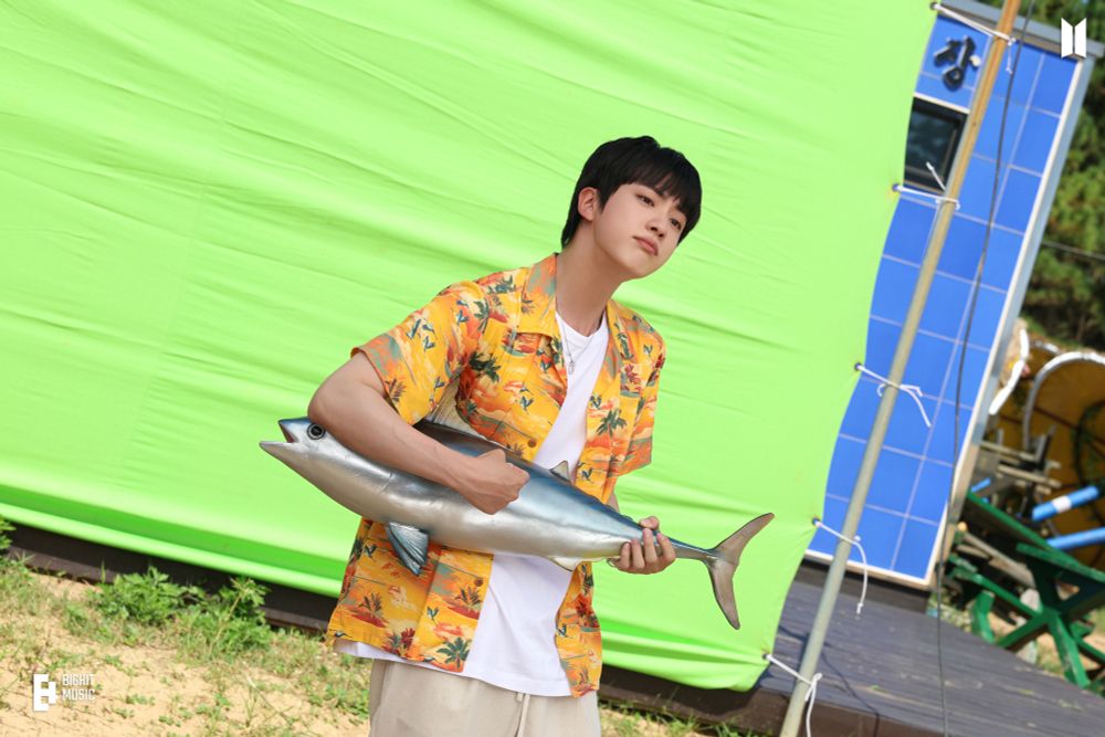 Jin holding a fake fish like a guitar while wearing a yellow/ orange shirt in front of a green screen 