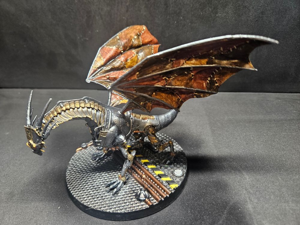 A miniature mechanical dragon with leather wings sits on a base that appears to be the floor of a factory.  