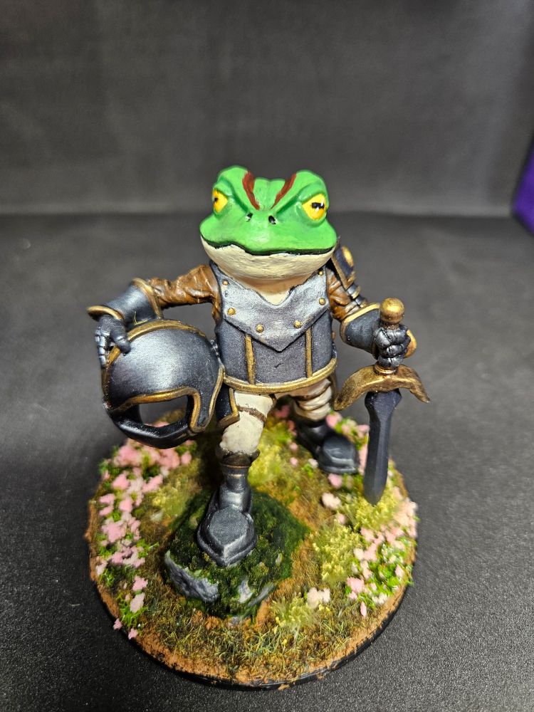 A miniature frog knight stands proudly with one foot atop a misty rock. He is surrounded by pink flowers. 