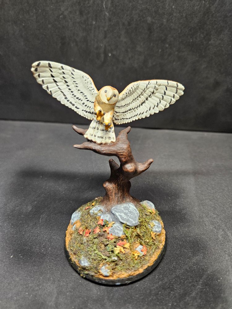 A miniature owl swoops or if the air.  It's real feathers rest on a dead tree on top of a rocky and grassy base.  