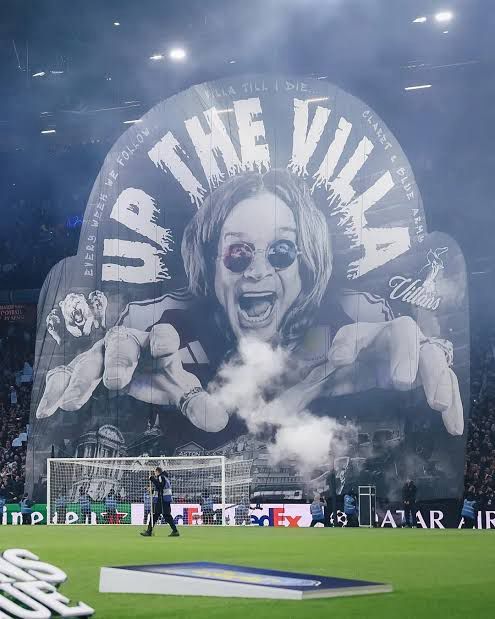 Torcida do Aston Villa levantando um bandeirão com imagem do Ozzy e a frase Up the Villa
