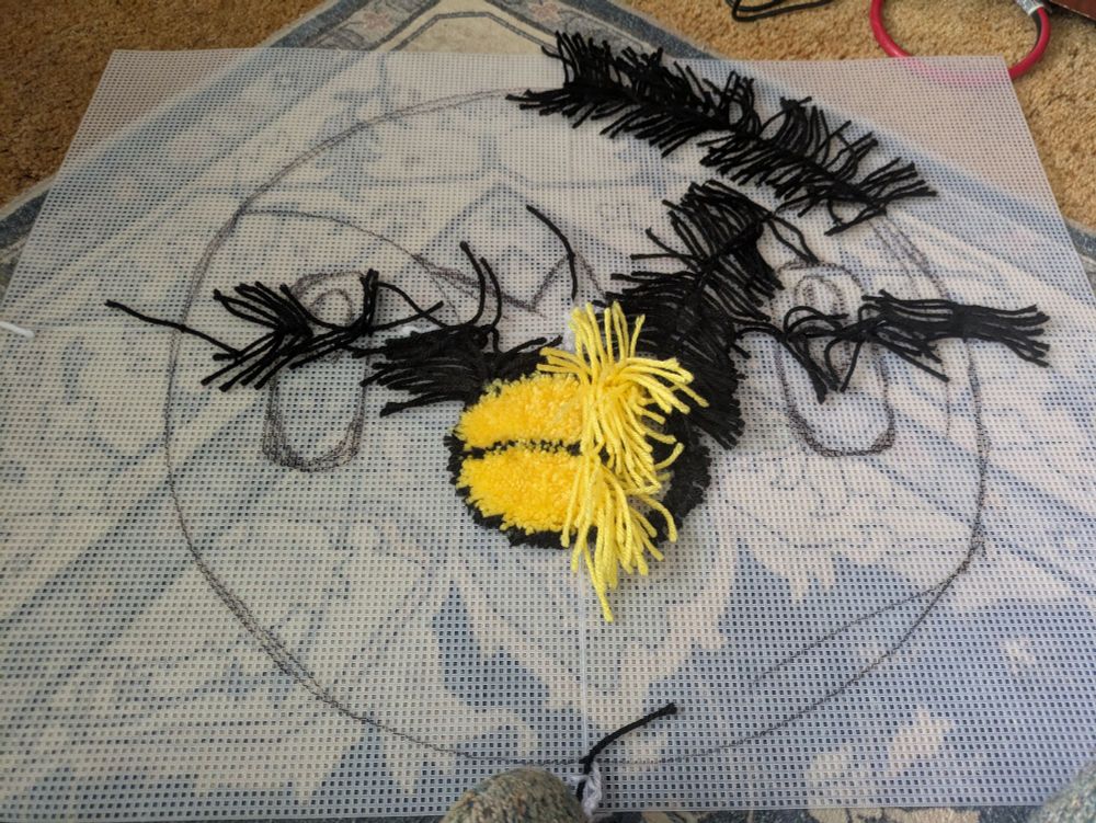 A plastic grid has the head of a Piplup drawn on it. Black yarn is in an outline to form the shape of a break. Yellow yarn fills the left side of the beak. Black yarn covers some parts of the Piplup head outline.