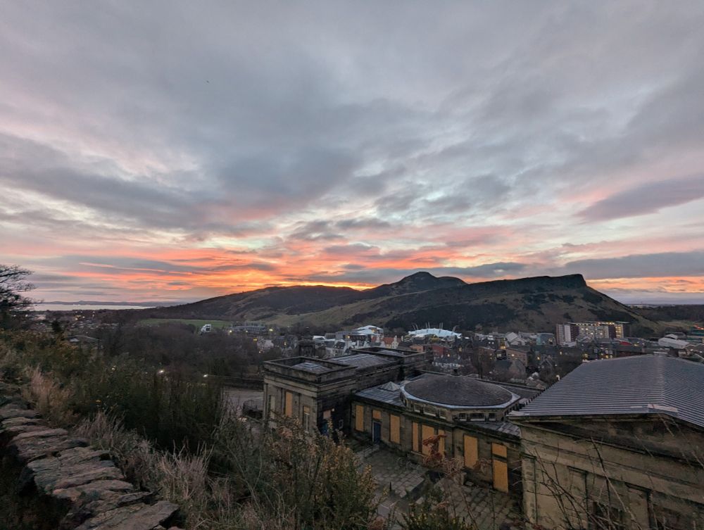 A view of a cloudy sky with a band of orange and red light from the sunrise over Arthur's Seat, a rocky hill in Edinburgh. In the foreground there are a range of buildings of differing styles and ages