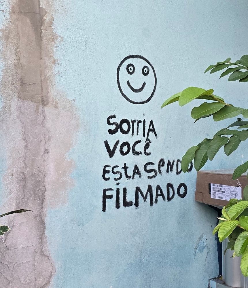Na foto tem uma parede e um aviso, sorria você está sendo filmado, com um rosto sorriso e com os olhos trincados