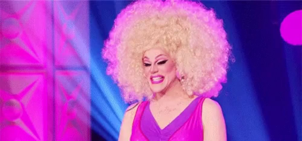 a drag queen wearing a large blonde afro wig and a pink dress is smiling .