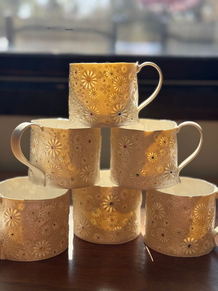 A stack of translucent porcelain mugs. The mugs are stamped with daisies, and the afternoon light is is shining through. 