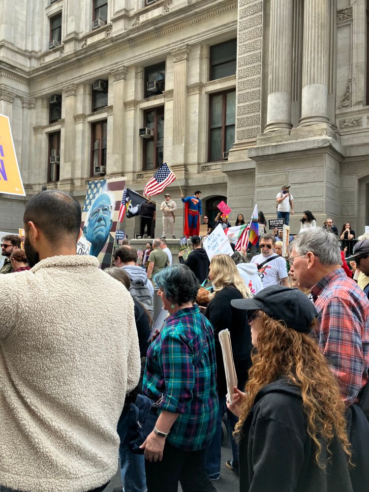 Marchers at Philadelphia No Kings rally. A man dressed as Superman is standing in the background waving a large American flag.