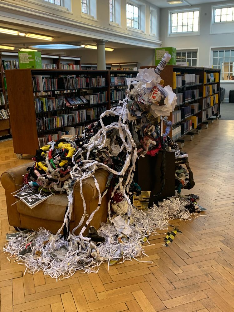 An arm chair covered in knotted tangled mess of paper forms that spill out over the floor. Surrounded by shredded paper tea bags an old mug 