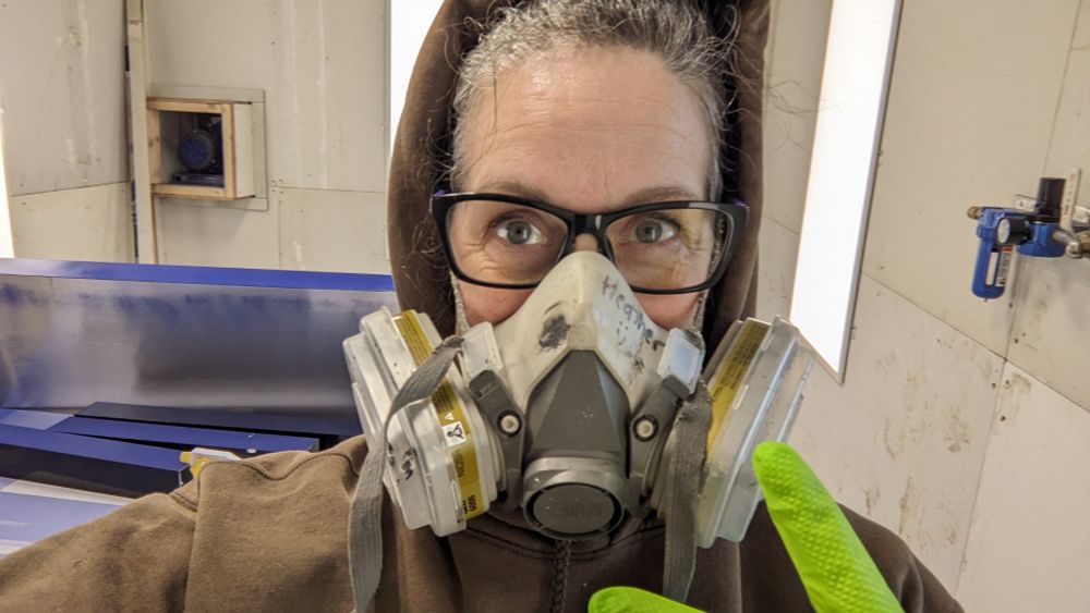 Grey haired woman in a paint booth wearing gloves and mask. 