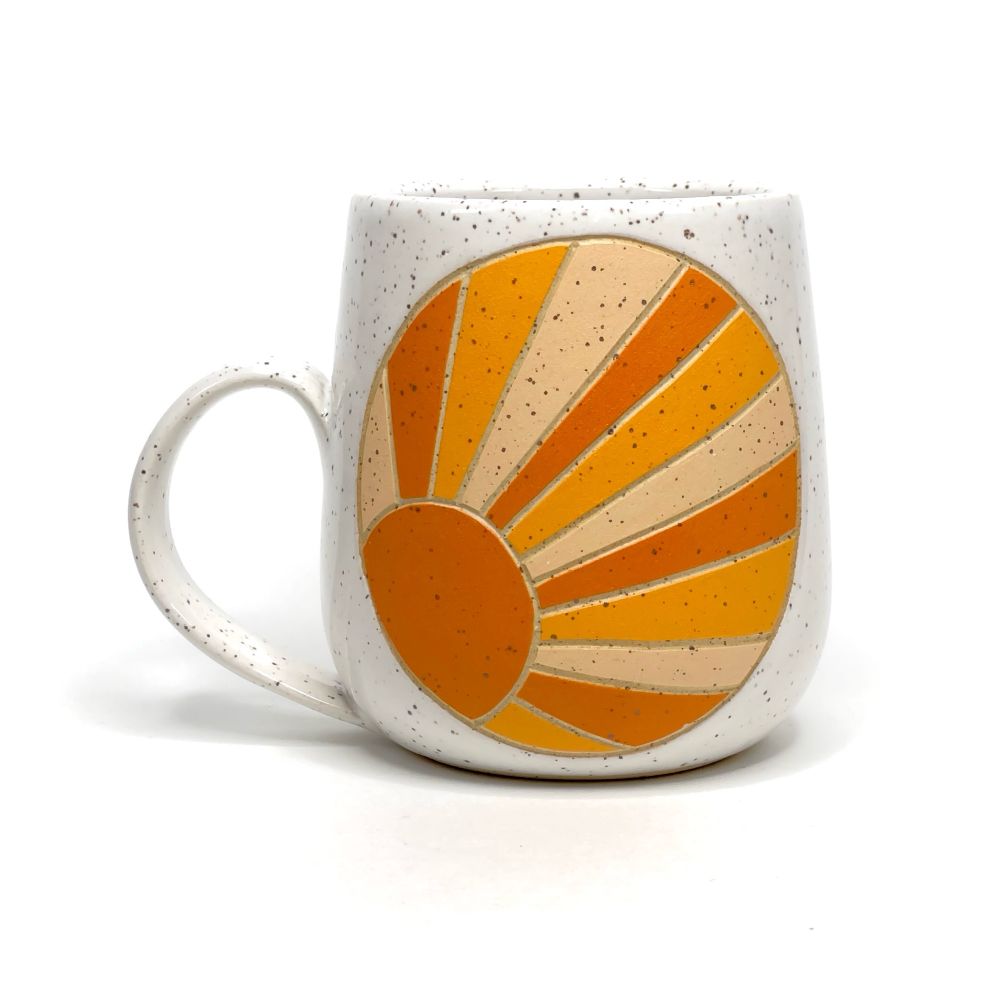 A white speckled mug with a carved illustration of an orange sun with dark, medium and bright orange rays, enclosed in a circle. 