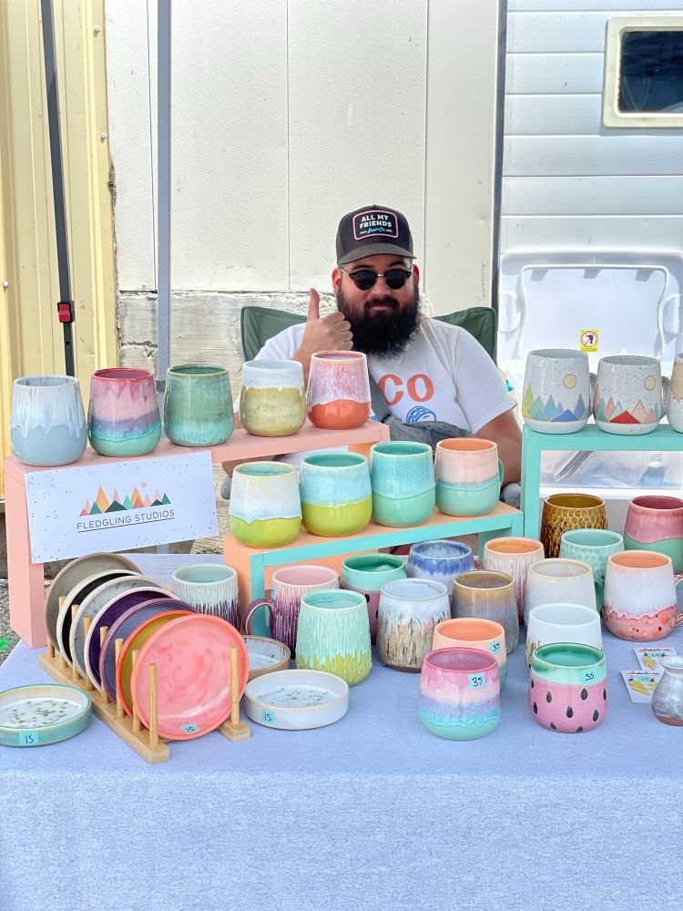 A smiling, dark haired bearded man with a black baseball hat and sunglasses giving a thumbs up as he sits behind a table with a large display of brightly coloured pottery. 