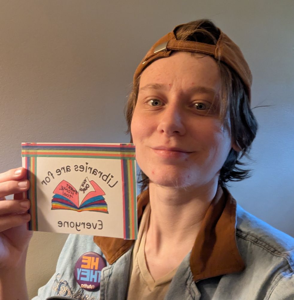 Carson holds up a postcard which reads "Libraries are For Everyone" and features a handwritten "This is Our Sweet Home, Too!" note within a rainbow paged book, to fight attempts to censor libraries in Alabama. 