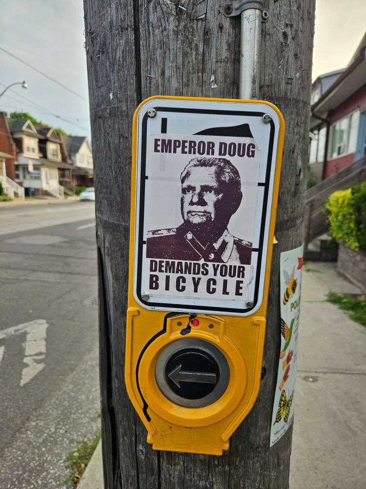Sticker on wooden telephone pole above  crosswalk button says Emperor Doug Demands Your Bicycle