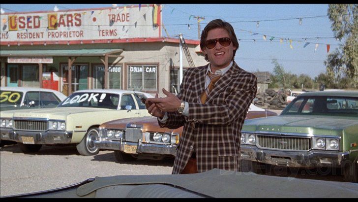 A smiling Kurt Russell dressed in sunglasses, a plaid jacket, and ill-fitting yellow tie,  in front of a line of dilapidated used cars at a seedy dealership in the 1980 movie "Used Cars"