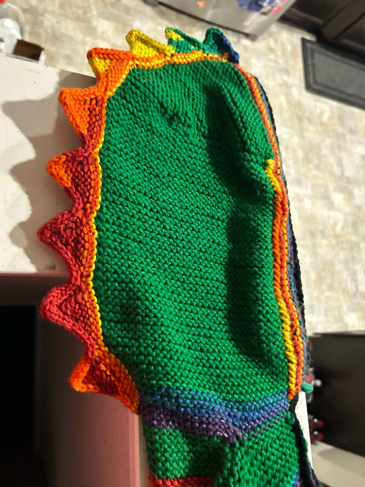 A green knitted hood with a sawtooth set of triangles as mostly orange dinosaur (stegosaur?) plates.