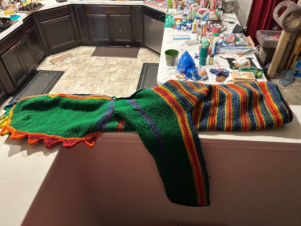 A hooded sweater in green and variegated rainbow yarn, with long sleeves and a rainbow sawtooth border on the green hood. 