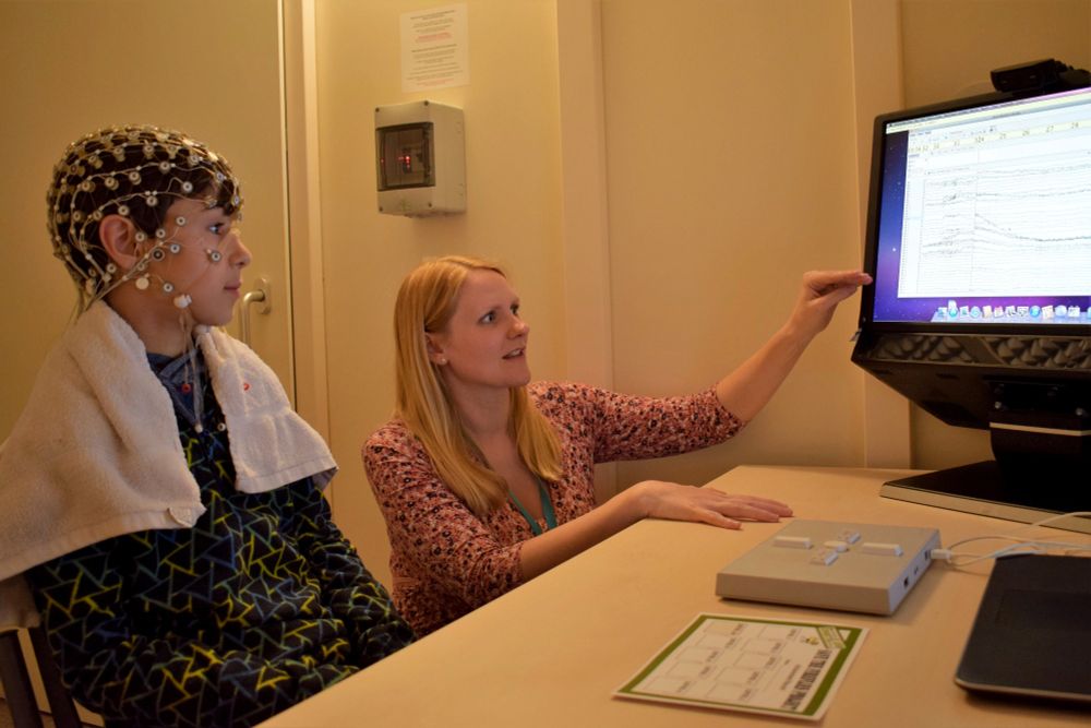 A child wearing a cap with wires to measure brain responses sits next to Dr Cathy Manning and looks at a computer screen.