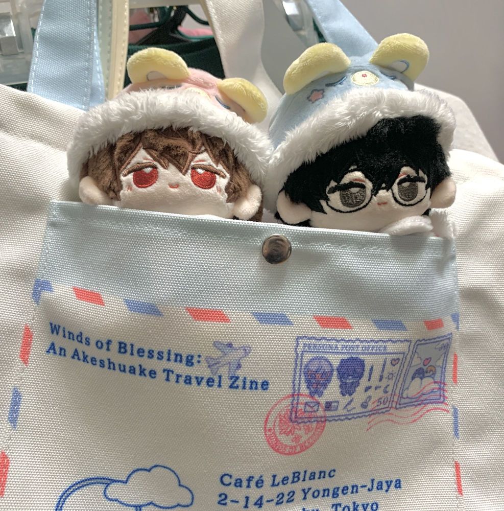 Pictured: Wind of Blessings: Akeshuake Travel Zine Tote bag holding two 10cm plushies of the P5 protagonist and Goro Akechi